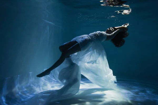 Iluminación en elegante mujer joven en vestido elegante blanco nadando en la piscina - foto de stock