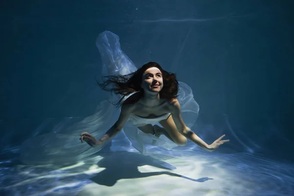 Iluminación en mujer joven feliz en vestido elegante blanco nadando en la piscina - foto de stock