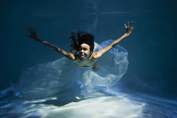 Освітлення на усміхненій молодій жінці в білому елегантному платті, що плаває в басейні — стокове фото