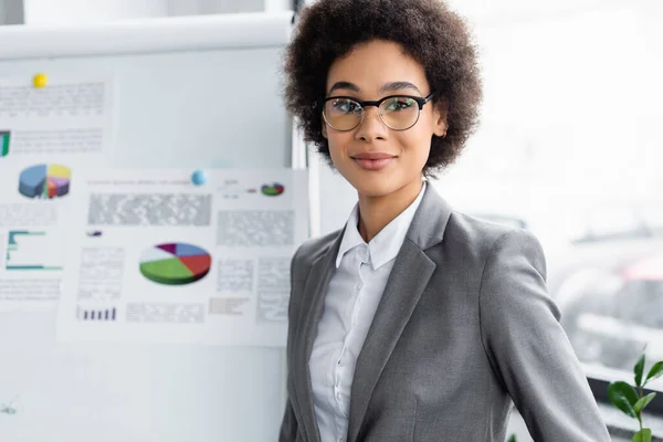 Joven afroamericano manager en traje mirando cámara cerca borrosa flipchart - foto de stock