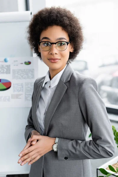 Gestionnaire afro-américain en costume et lunettes regardant la caméra près de tableau à feuilles mobiles — Photo de stock