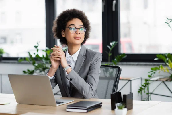 African american manager in eyeglasses looking away near laptop and notebooks in office — Stock Photo