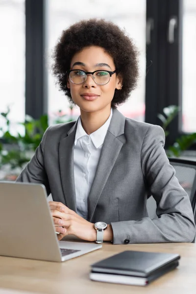 Afroamerikanische Geschäftsfrau in offizieller Kleidung blickt in die Kamera in der Nähe eines verschwommenen Laptops — Stockfoto