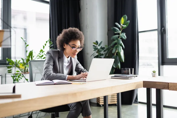 Young african american businesswoman in eyeglasses using laptop near notebooks on table — Stock Photo