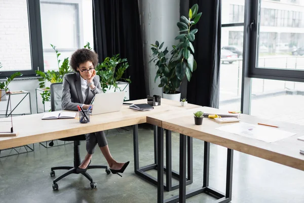 African american manager in eyeglasses looking at laptop near stationery on table — Stock Photo