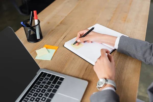 Vista recortada de la mujer de negocios escribiendo en nota adhesiva en el cuaderno cerca de la computadora portátil borrosa — Stock Photo