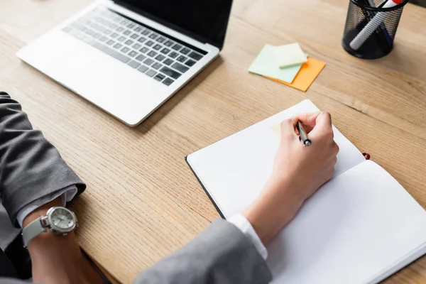 Обрізаний вид бізнес-леді в офіційному носінні, що пише на блокноті біля розмитого ноутбука на столі — стокове фото