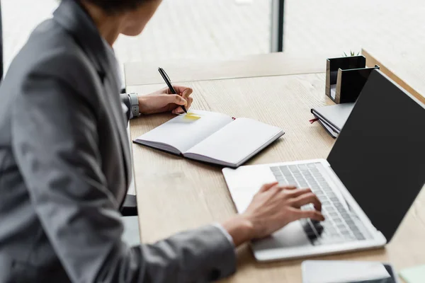 Cropped view of african american businesswoman writing on notebook and using laptop with blank screen — Stock Photo