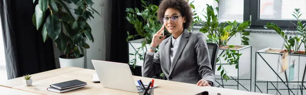 Smiling african american businesswoman talking on smartphone near laptop on table, banner — Stock Photo