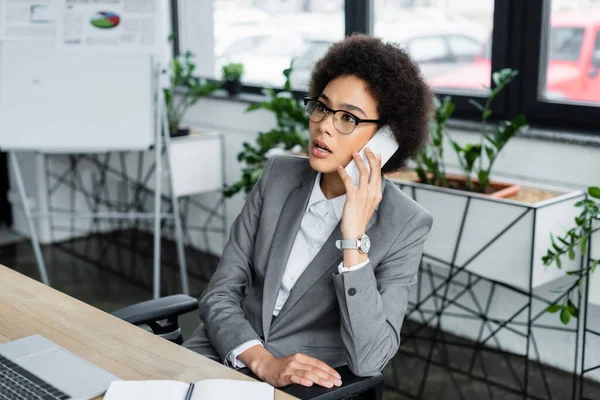 African american manager talking on smartphone near blurred laptop and notebook — Stock Photo