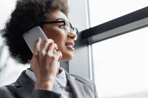 Vista de ángulo bajo del gerente afroamericano positivo hablando en el teléfono inteligente cerca de la ventana - foto de stock