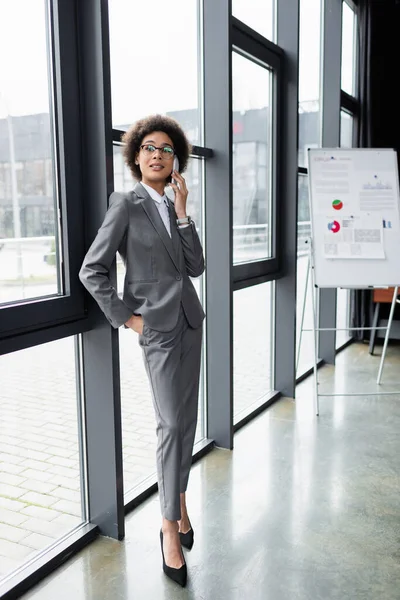 Empresária afro-americana positiva falando no celular perto da janela no escritório — Fotografia de Stock