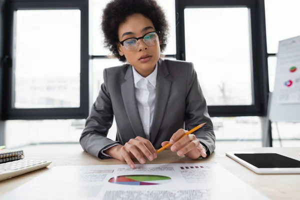 Femme d'affaires afro-américaine en lunettes tenant un crayon près du papier avec des graphiques et une tablette numérique — Photo de stock