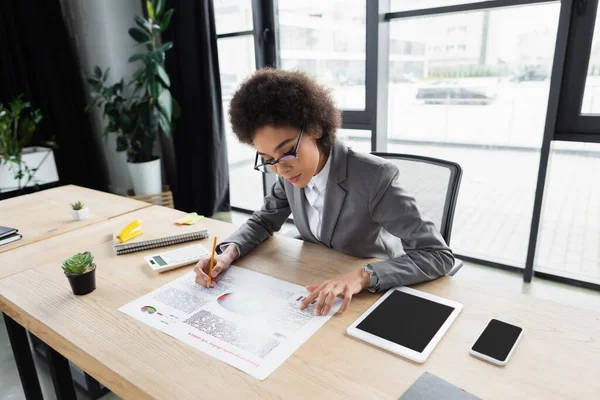 Gerente afroamericano escribiendo en documento cerca de dispositivos con pantalla en blanco - foto de stock