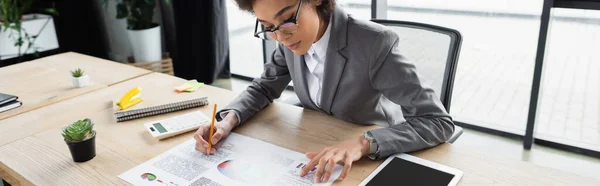 Afroamerikanische Geschäftsfrau schreibt auf Papier mit Diagrammen in der Nähe von digitalem Tablet, Banner — Stockfoto