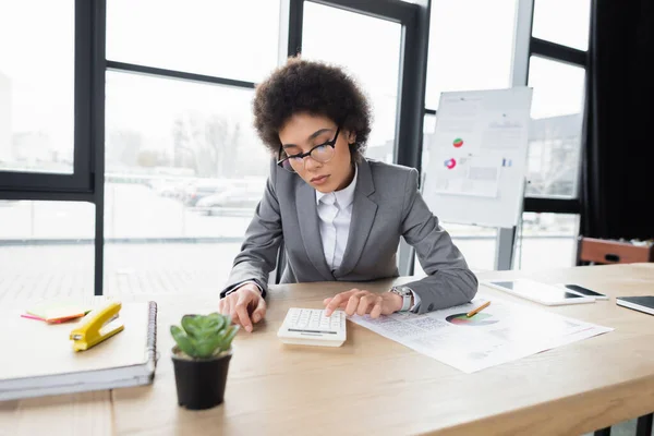 Afrikanischer Manager arbeitet mit Taschenrechner in der Nähe digitaler Tablets und Notebooks — Stockfoto