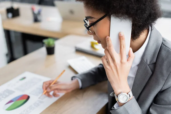 African american manager in eyeglasses talking on mobile phone and writing on blurred paper — Stock Photo