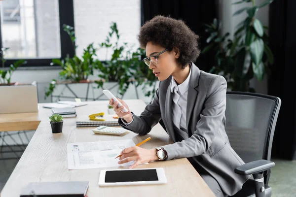 African american manager using smartphone and holding pencil near document and stationery — Stock Photo