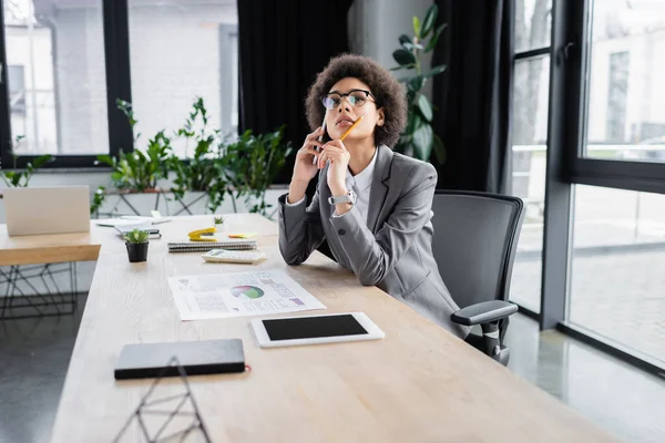 Pensativa mujer de negocios afroamericana con lápiz hablando en el teléfono inteligente en la oficina - foto de stock