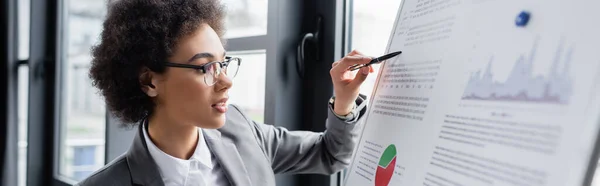 Junger afrikanisch-amerikanischer Manager hält Stift in der Nähe von Dokumenten auf verschwommenem Flipchart, Banner — Stockfoto