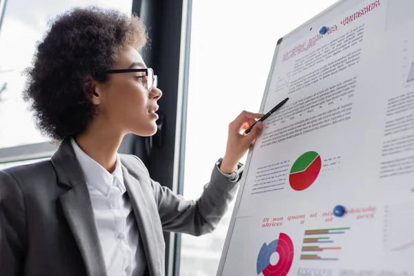 Vista lateral de una mujer de negocios afroamericana con gafas que sostiene la pluma cerca del documento con gráficos - foto de stock