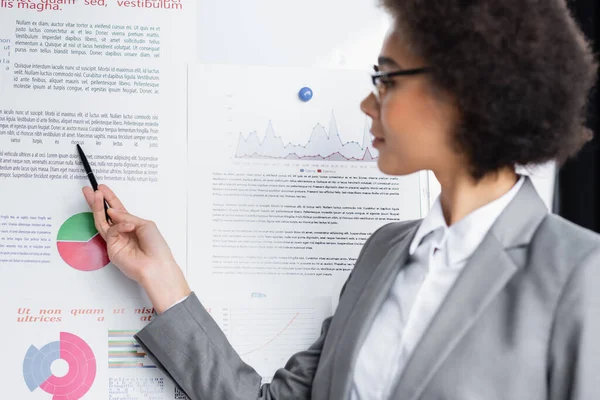 Femme d'affaires afro-américaine floue pointant vers un tableau à feuilles tout en tenant un stylo — Photo de stock