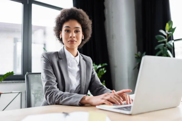 African american businesswoman in earphones looking at camera near blurred laptop — Stock Photo
