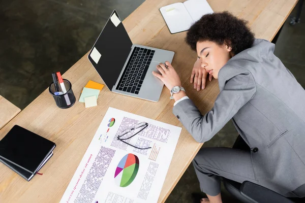 Hochwinkelaufnahme einer afrikanisch-amerikanischen Geschäftsfrau, die mit geschlossenen Augen in der Nähe von Laptop und Papier auf dem Tisch liegt — Stockfoto