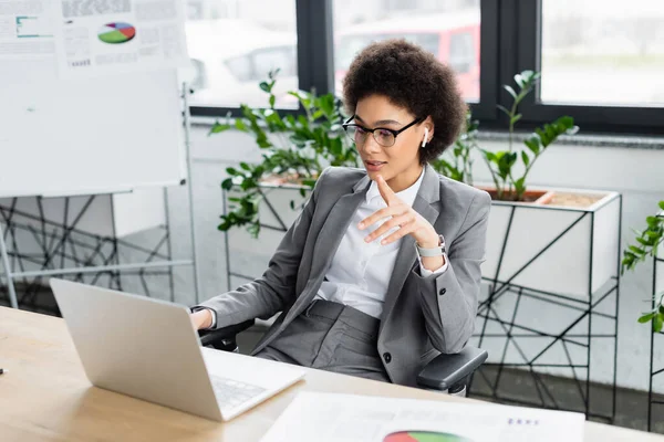 Empresária afro-americana em fone de ouvido usando laptop e apontando com o dedo no escritório — Fotografia de Stock