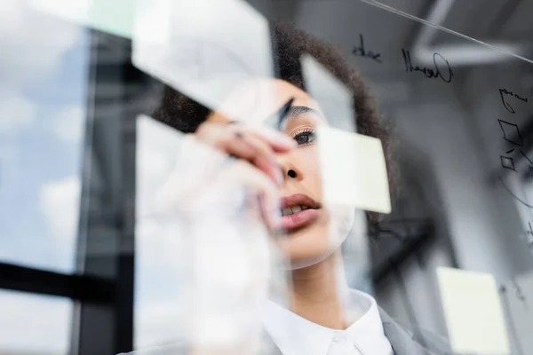 Vista de ángulo bajo de la mujer de negocios afroamericana sosteniendo marcador borroso cerca de tablero de vidrio - foto de stock