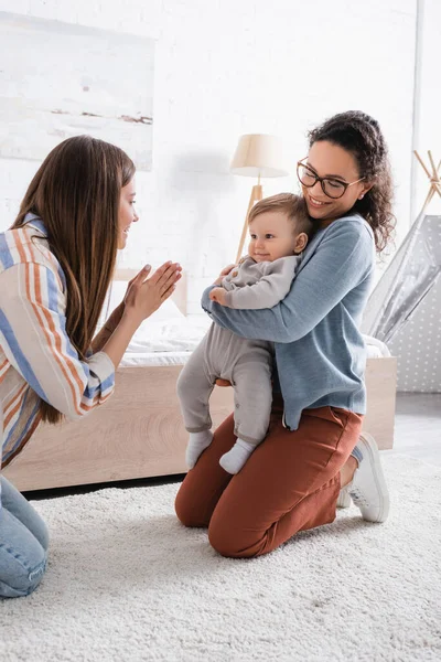 Lächelnde Afroamerikanerin mit Brille, die einen kleinen Jungen in der Nähe der fürsorglichen Mutter hält und in die Hände klatscht — Stockfoto