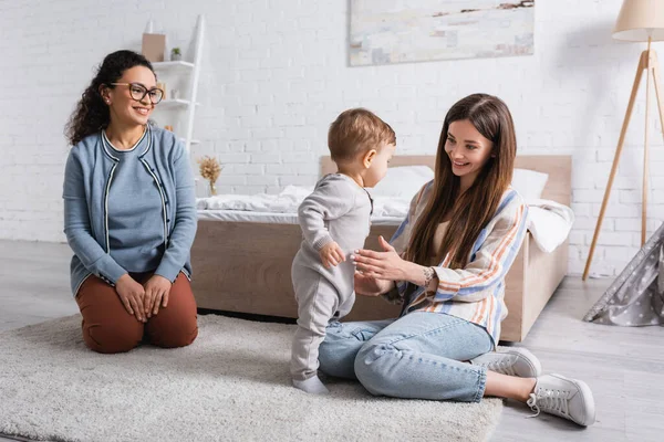 Feliz interracial mulheres olhando para bebê menino de pé no tapete — Fotografia de Stock