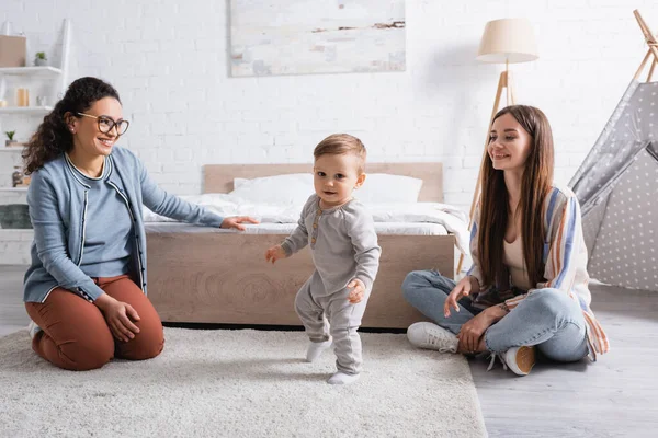 Cheerful interracial women looking at baby boy walking on carpet — Stock Photo