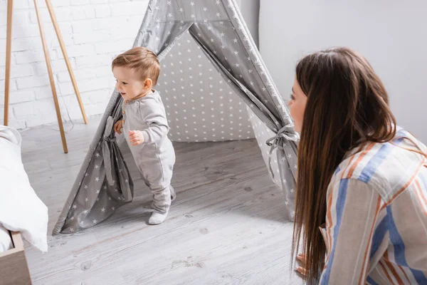 Mãe desfocada olhando para o bebê filho satisfeito de pé perto de tipi no quarto — Fotografia de Stock