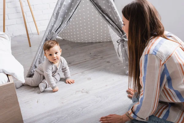 Blurred mother looking at pleased baby son sitting near tipi in bedroom — Stock Photo