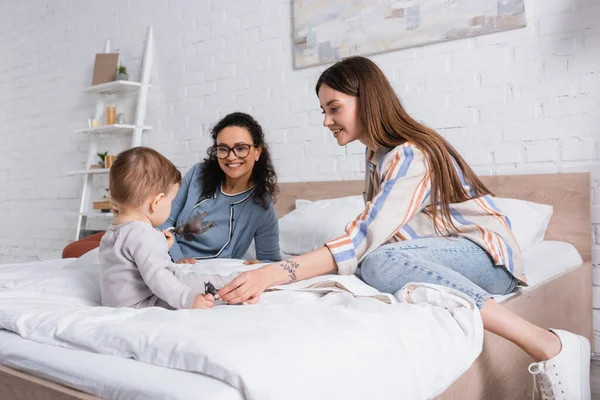 Happy tattooed mother looking at infant son while sitting on bed near african american woman — Stock Photo