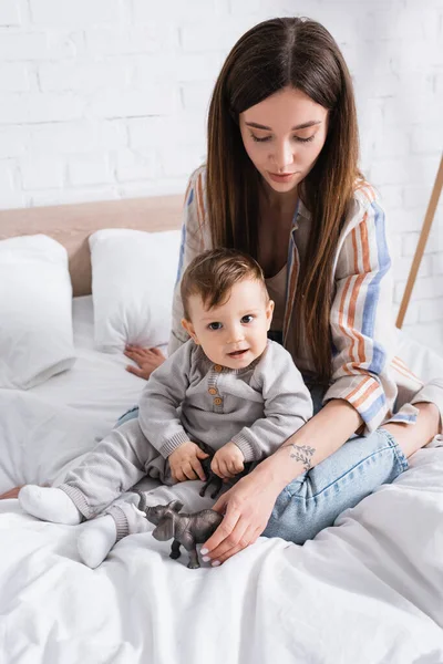 Tattooed mother holding toy near infant son while sitting on bed — Stock Photo