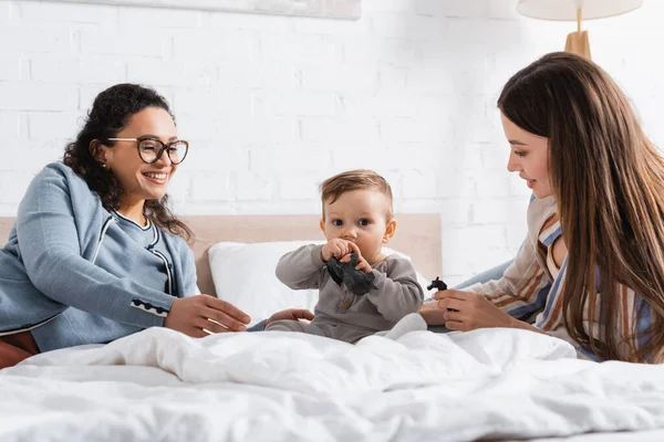 Feliz interracial mulheres olhando infantil menino brincando com brinquedo enquanto sentado na cama — Fotografia de Stock