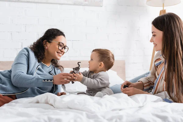 Alegre interracial mulheres olhando para infantil menino brincando com brinquedo enquanto sentado no cama — Fotografia de Stock