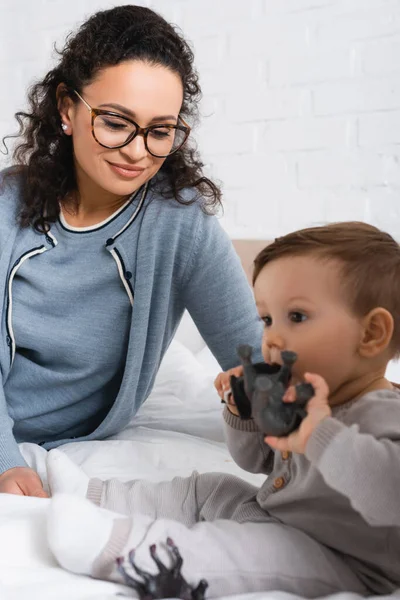 Happy african american woman looking at blurred infant boy playing with toy on bed — Stock Photo