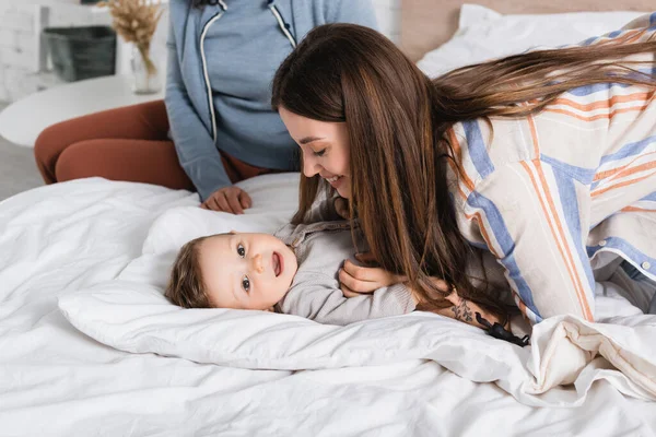 Glückliche Mutter kitzelt Säugling neben Frau, die auf Bett sitzt — Stockfoto