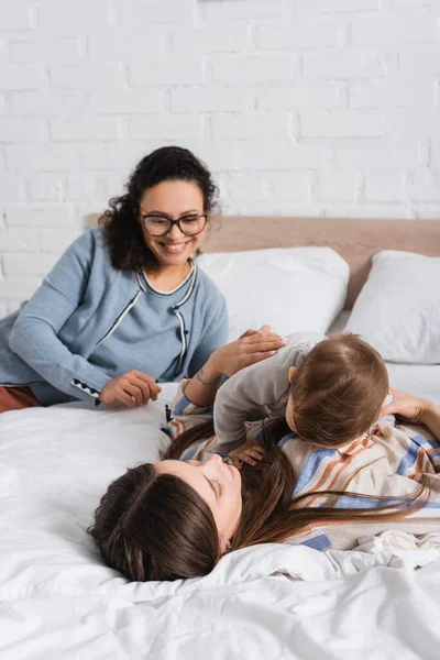 Mother and infant boy lying on bed near blurred african american woman in glasses — Stock Photo