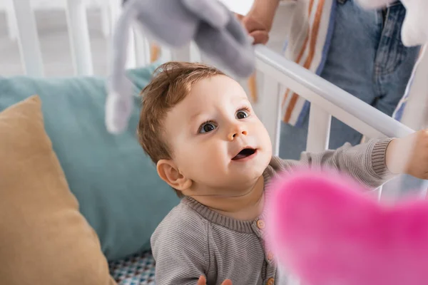 Menino infantil espantado no berço do bebê com brinquedos macios pendurados em primeiro plano desfocado — Fotografia de Stock