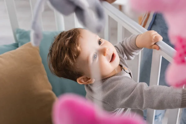 Menino no berço com brinquedos macios pendurados em primeiro plano borrado — Fotografia de Stock