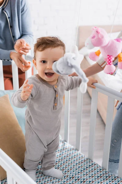 Excited infant boy in baby crib with hanging soft toys on blurred foreground — Stock Photo