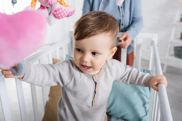 Menino infantil alegre no berço do bebê com brinquedo macio pendurado em primeiro plano desfocado — Fotografia de Stock