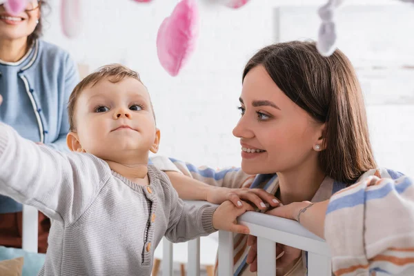 Усміхнена мати дивиться на сина, який досягає м'яких іграшок, що звисають над ліжком — стокове фото