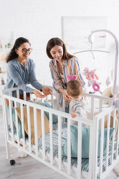 Happy interracial women looking at infant boy in baby crib at home — Stock Photo