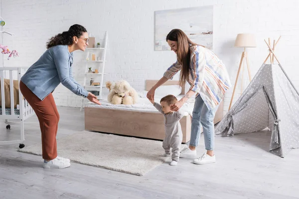 Mother supporting baby boy walking near smiling african american woman in glasses — Stock Photo