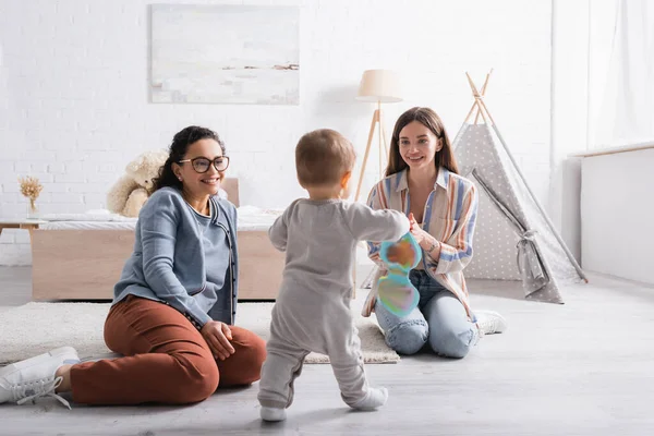 Niño pequeño caminando con libro de bebé cerca de mujeres interracial en el dormitorio - foto de stock